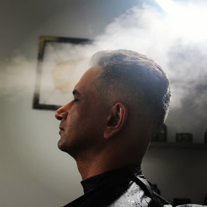A client in the chair with eyes closed, warm steam rising around a fresh fade